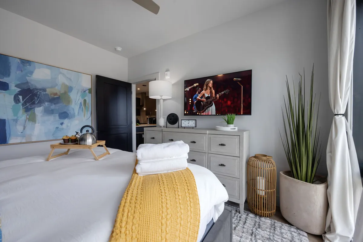 A stylish bedroom featuring a comfortable bed dressed in crisp white linens and a yellow throw, a large abstract painting above the headboard, a TV displaying entertainment, a potted plant, and a nightstand with a lamp. The room exudes comfort and offers amenities for relaxation and entertainment.