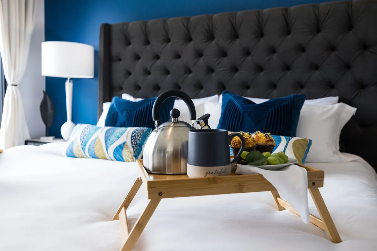 A cozy bedroom with a tufted headboard, decorative pillows, a breakfast tray on the bed, and a contemporary light fixture. Soft lighting and elegant touches create a luxurious yet welcoming ambiance.