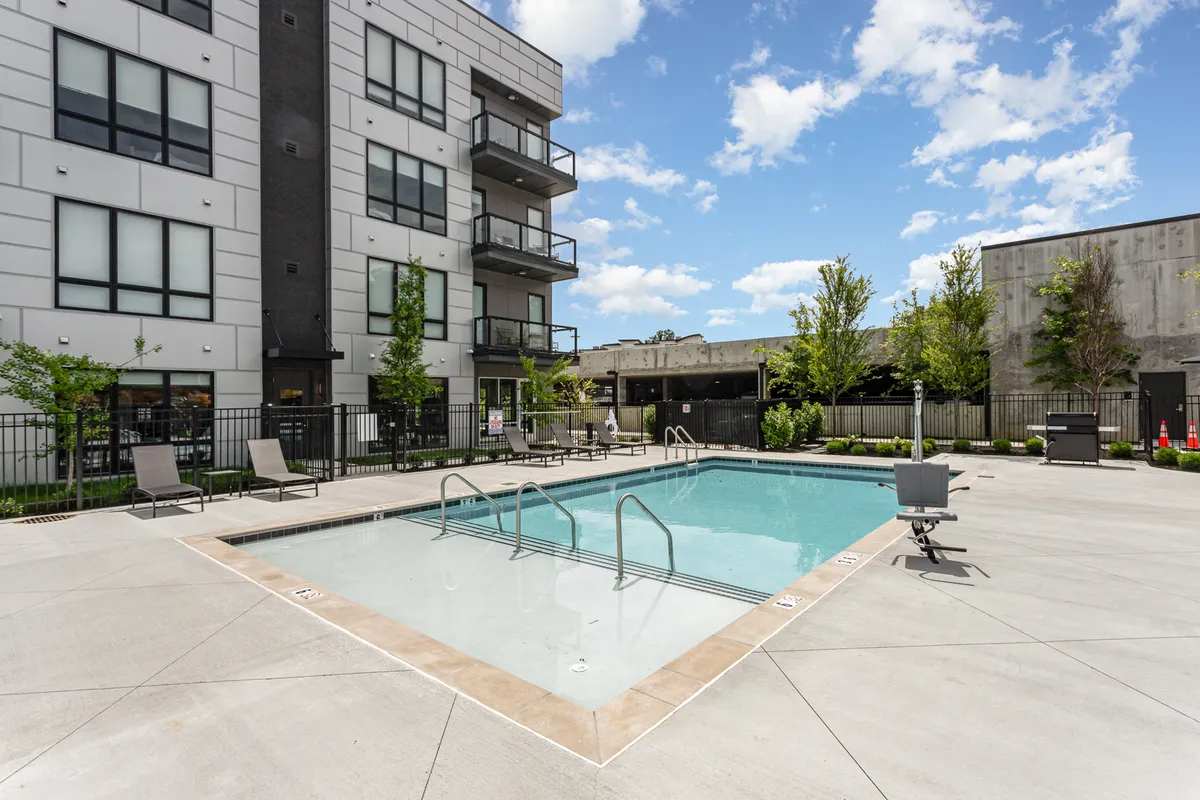 A wide-angle exterior shot of a swimming pool with inviting lounge chairs, balconies above, and verdant greenery. The scene radiates a refreshing and relaxing mood ideal for both quiet sunbathing and socializing with friends or family.