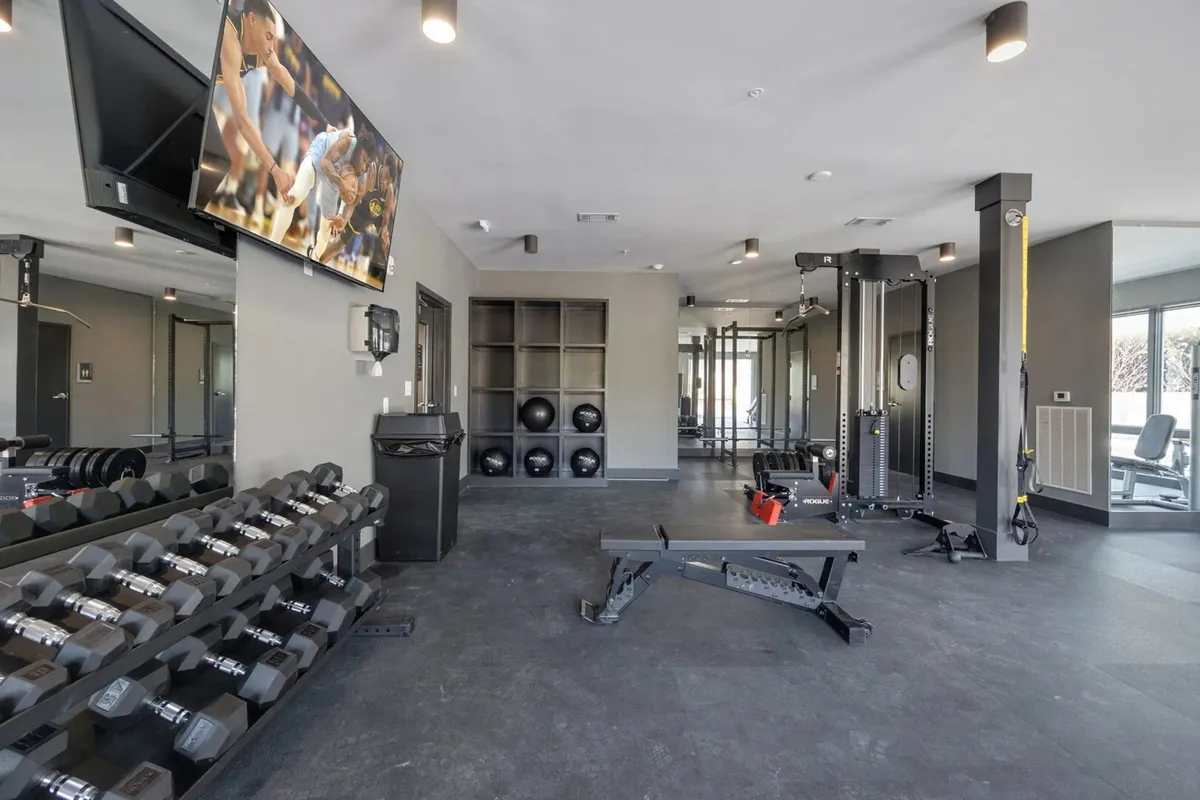 An energetic interior view of a modern fitness center featuring a dumbbell rack, weight bench, exercise machines, a large television, and a mirrored wall. The environment is lively, well-equipped, and perfect for a range of fitness activities.