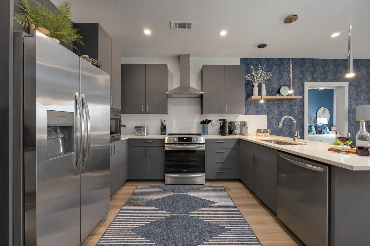A vibrant, contemporary kitchen with stainless steel appliances, a colorful backsplash, decorative plants, and a dining setup on the counter. The scene captures a sense of activity with cooking and casual dining, while bright lighting and stylish accents create a cheerful atmosphere.