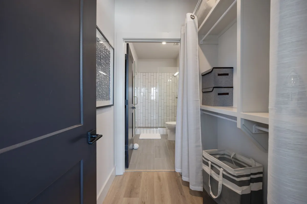 A fresh, inviting view with an open door leading to a bathroom, neatly arranged storage bins, and textured wall art. The image emphasizes organization, convenience, and a smooth transition between bedroom and bath spaces.