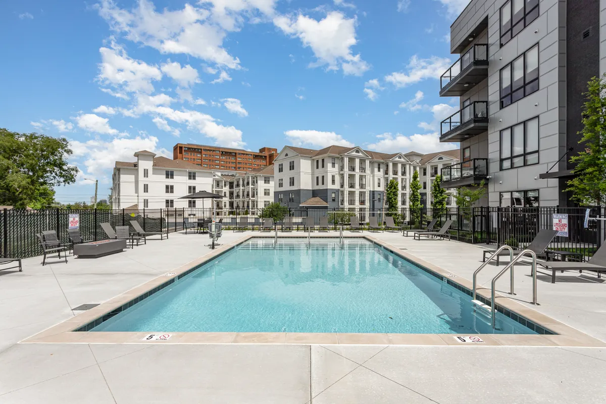 A resort-style swimming pool surrounded by lounge chairs and greenery, framed by modern buildings. The setting is tranquil yet luxurious, ideal for sunbathing, relaxing, or enjoying a quiet moment together.