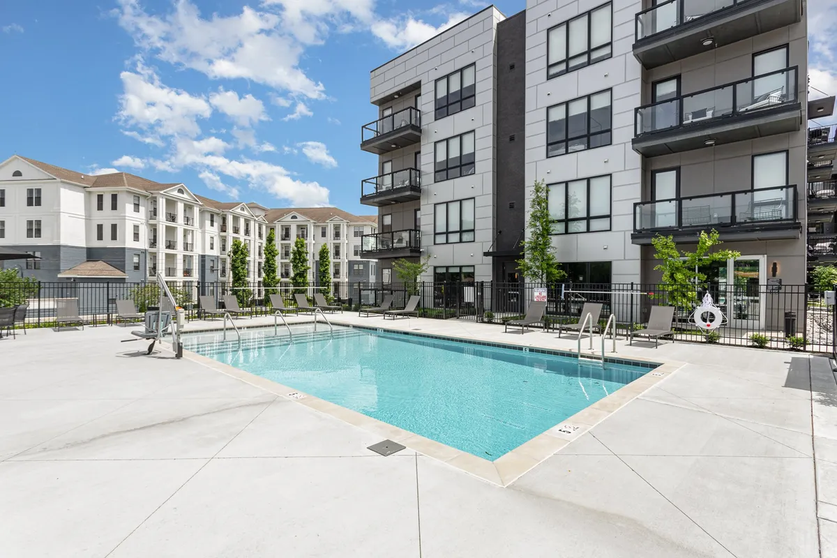 This exterior image showcases a sparkling swimming pool surrounded by modern apartment buildings and lush, landscaped greenery. Lounge chairs line the poolside, inviting residents to relax and socialize in a vibrant, resort-like setting.